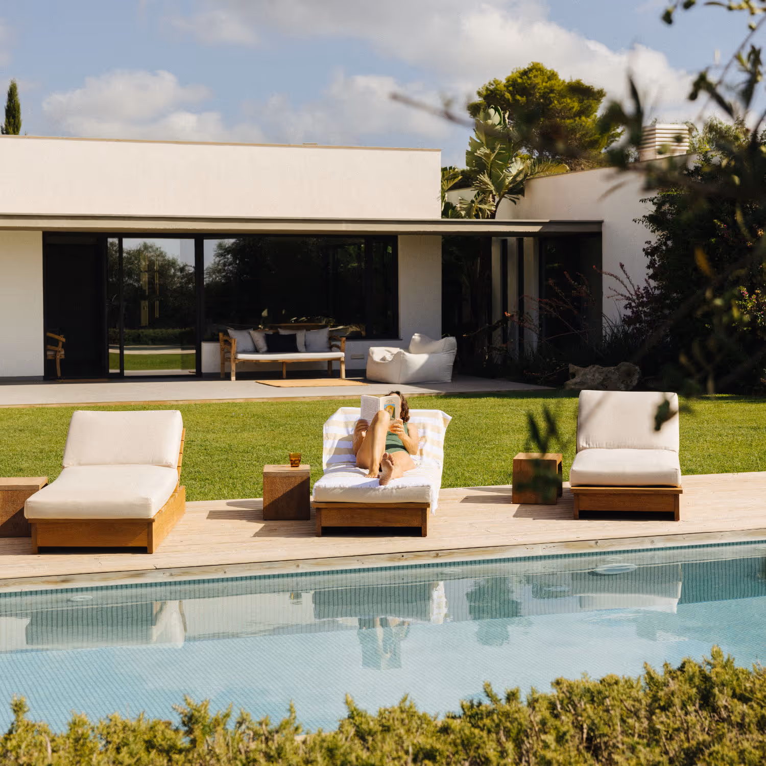 Services, discover your new property. The image shows a modern house in a rustic setting, with a swimming pool in the foreground and an exuberant grass field surrounded by Mediterranean plants. In the terrace there are 3 sunbeds, with a woman sunbathing in the central one whilst reading a book.