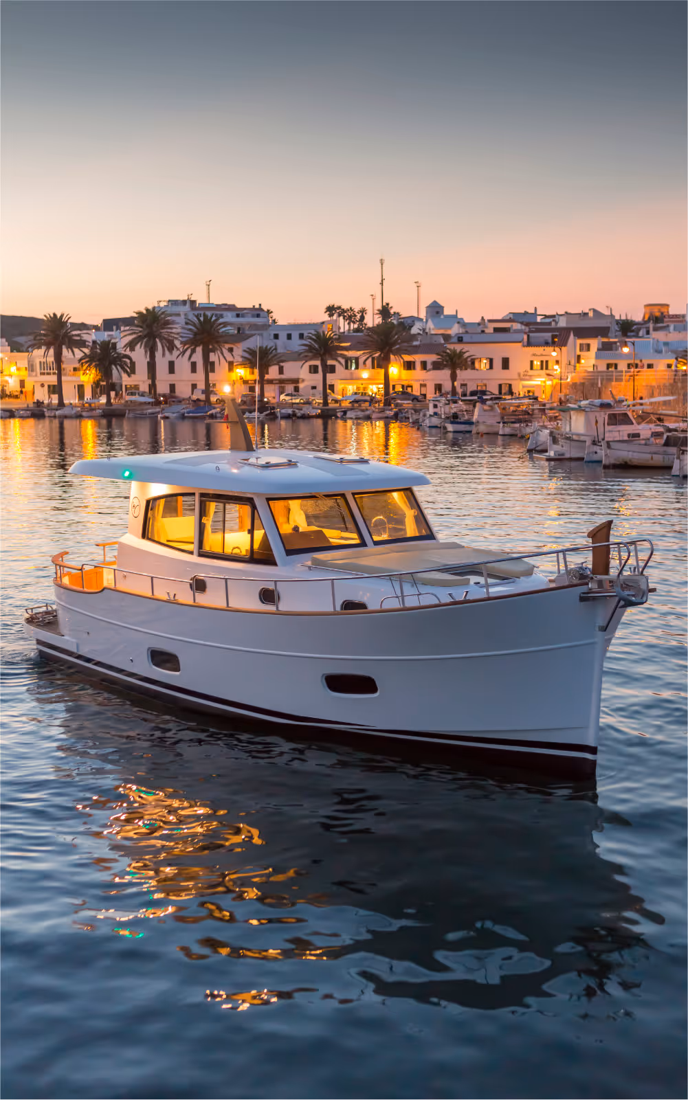Image that shows a Menorquin llaut yacht that is sailing the calm waters of Fornells bay. Fornells can be seen in the background with a beautiful sunset sky that gives a dreamy feel to the image.